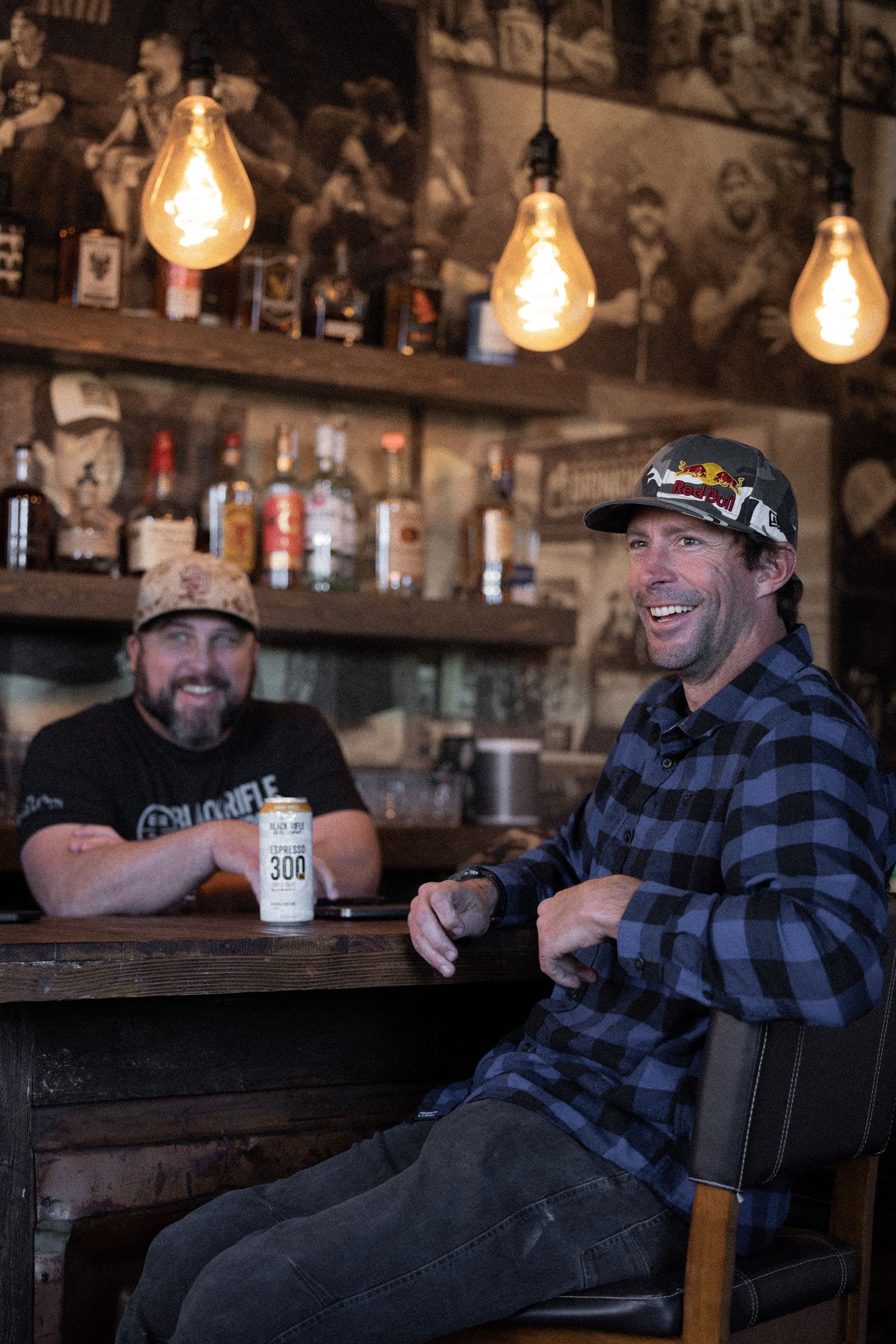 Two men talking at a wood bar under warm Edison bulbs, a coffee can on the counter
