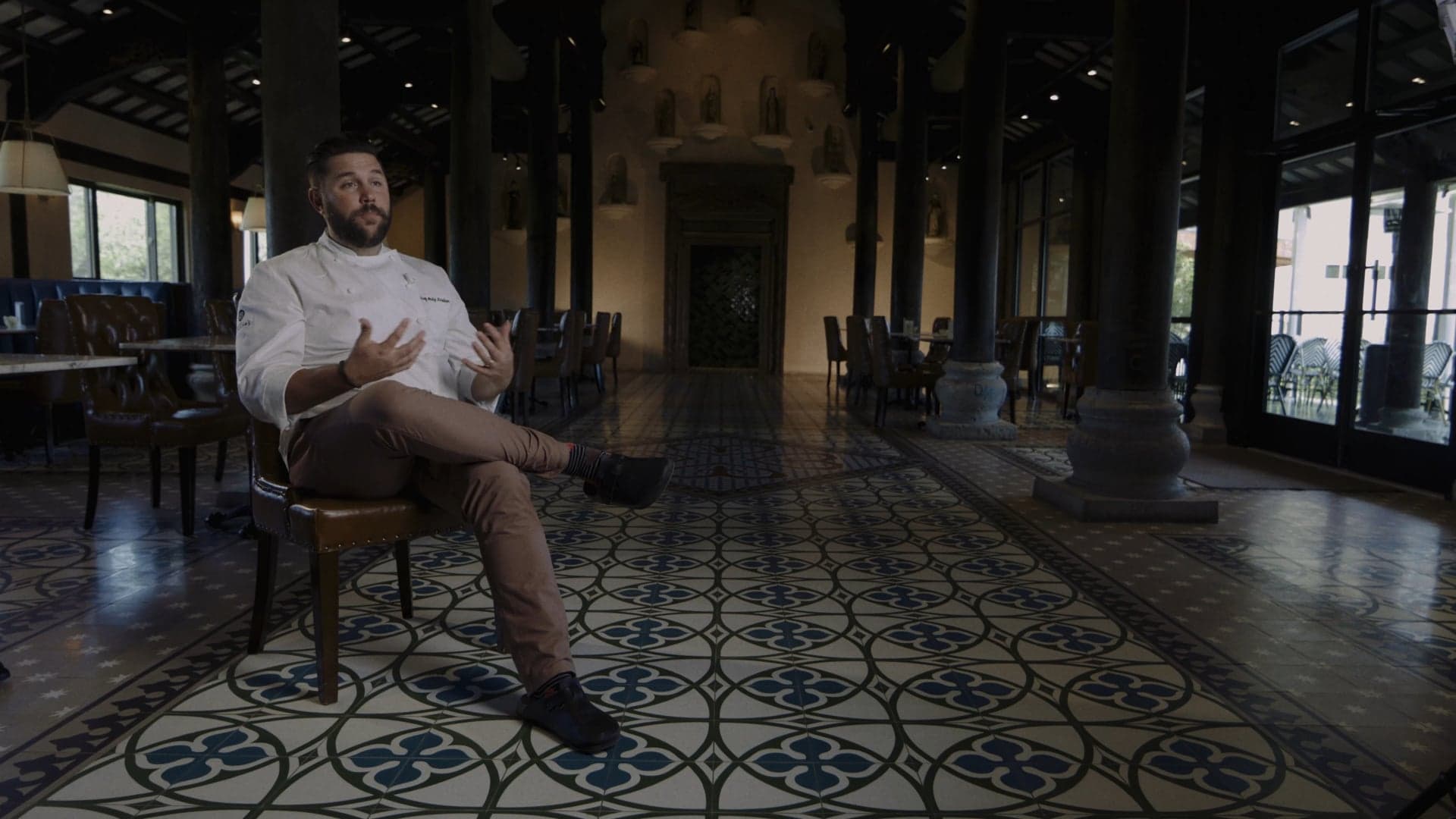 Chef in a white coat seated for an interview inside Tillie's Restaurant at Camp Lucy, with patterned tile floors and wooden pillars